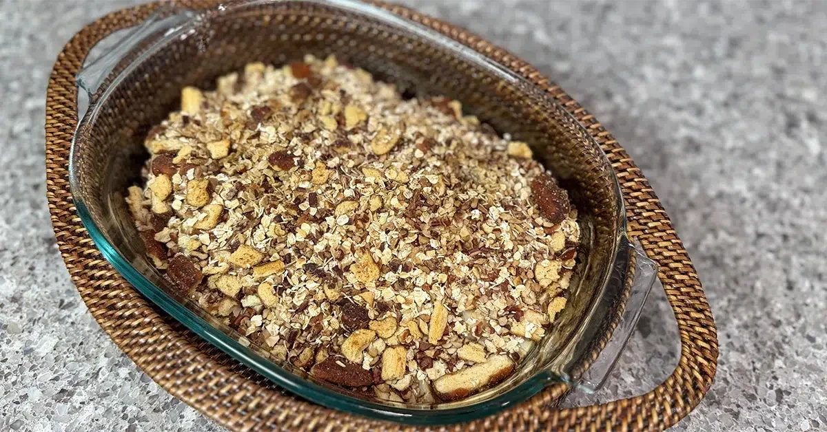 A glass bowl filled with a mixed nut and oat granola sits on a woven rattan tray atop a speckled gray countertop, conveying a rustic, homemade vibe.
