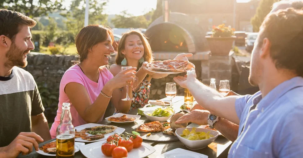 A group of people laughing and sharing a meal at a picnic table in a sunny outdoor setting.