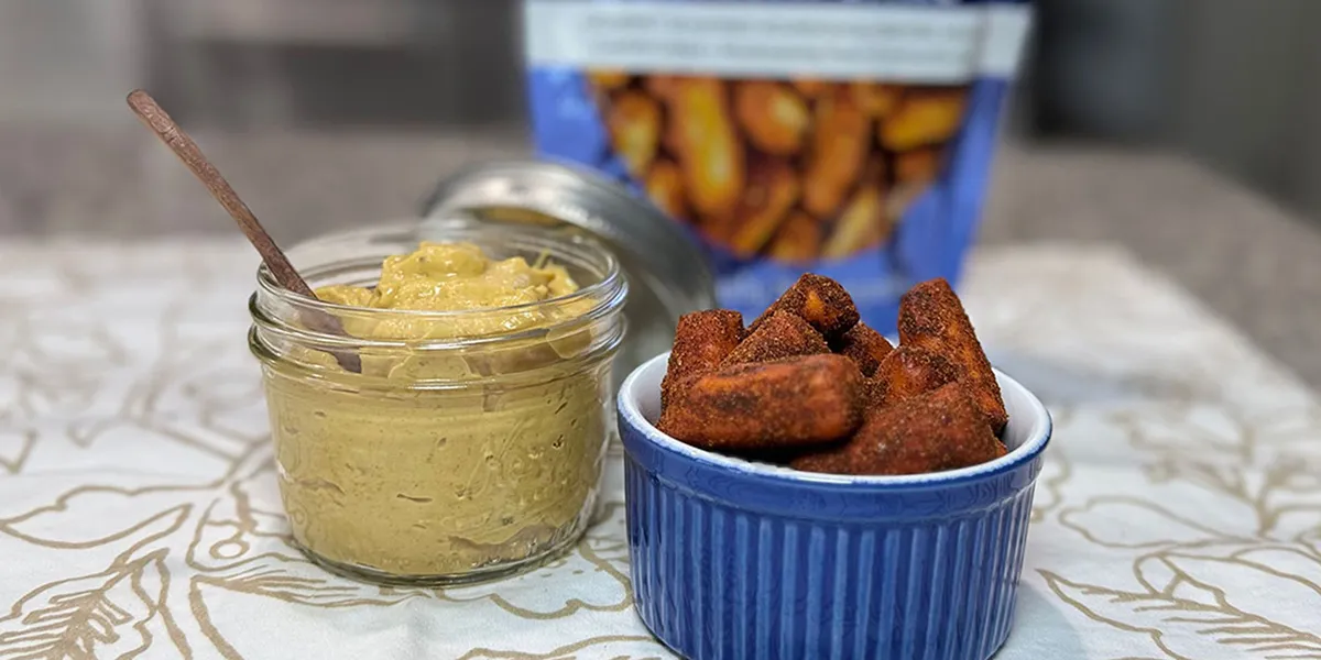 A jar of creamy hummus next to a bowl of golden, crispy fries on a wooden table.