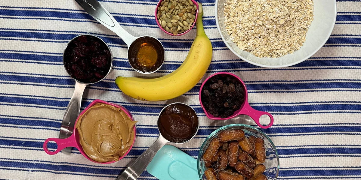Ingredients for a snack on a striped cloth: oats, banana, raisins, dried berries, seeds, honey, peanut butter, apple butter, and dates in measuring cups.