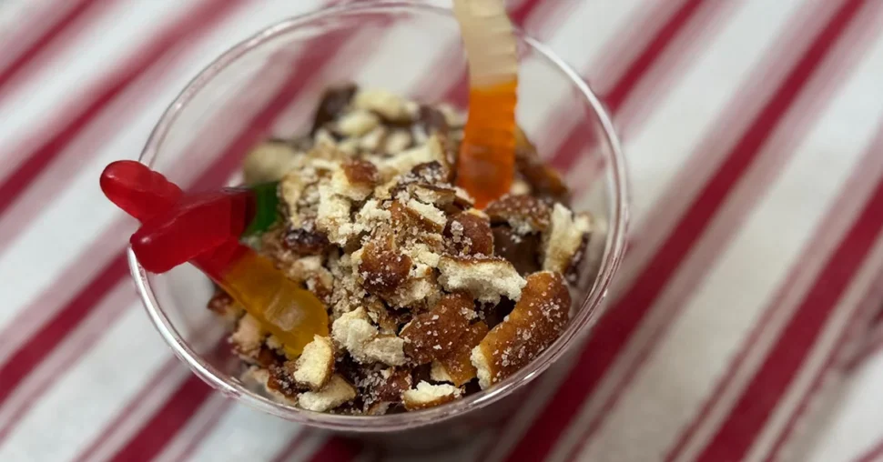 A cup filled with chocolate pudding topped with crumbled cookies and colorful gummy worms. Background features a red and white striped fabric.