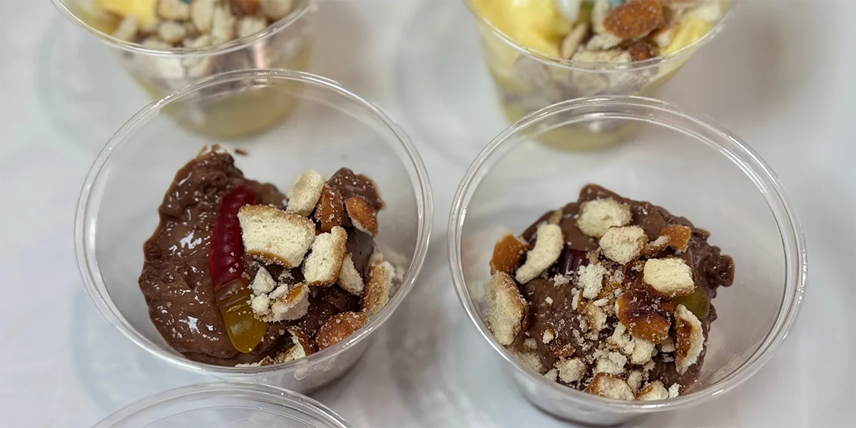 Plastic cups filled with chocolate pudding topped with crumbled cookies and a gummy worm on a light surface. The setting is casual and inviting.