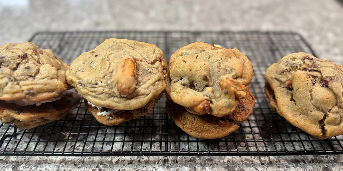 Freshly baked chocolate chip cookies with chunks of chocolate and caramel bits, neatly arranged in pairs on a wire rack on a granite countertop.