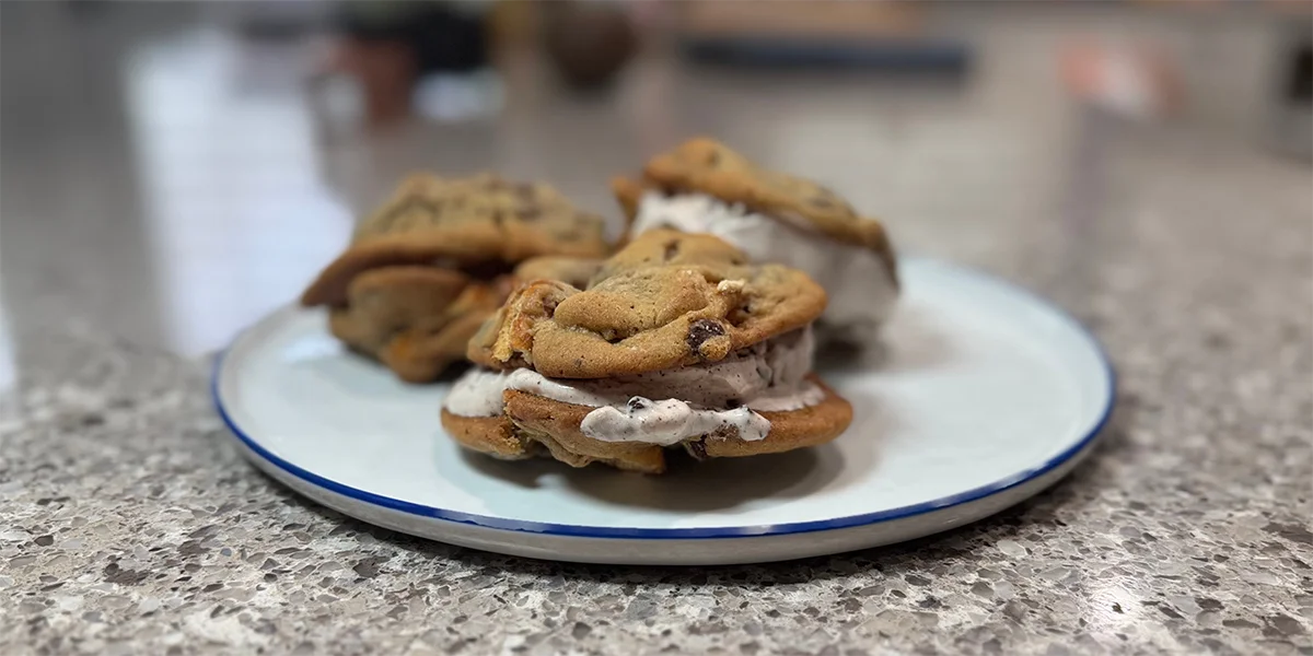 Three chocolate chip cookie ice cream sandwiches on a white and blue plate, resting on a speckled countertop, suggesting a sweet, homemade treat.