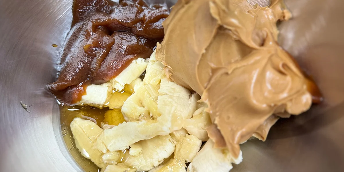 Close-up of a mixing bowl containing glossy brown applesauce, creamy peanut butter, sliced bananas, and honey. The tone is inviting and rich.