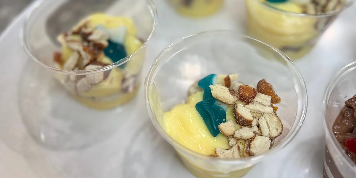 Close-up of a dessert cup with creamy yellow pudding, topped with crushed nuts and a blue gummy candy, on a white surface. Bright and inviting.
