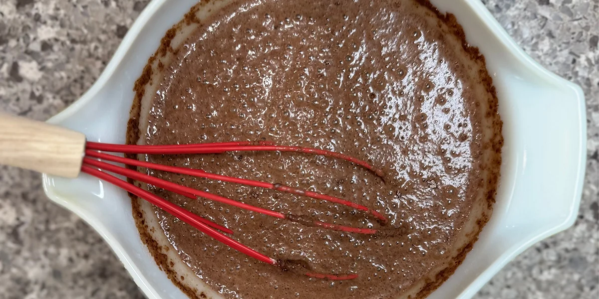 A white bowl on a speckled countertop holds a bubbly chocolate batter. A red whisk with a wooden handle is partially submerged, suggesting mixing.