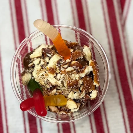 A glass cup with crumbled cookies and chocolate pudding topped with colorful gummy worms, sits on a red and white striped cloth, creating a playful dessert.