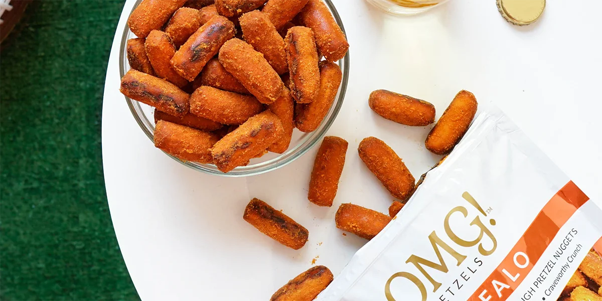 A bowl of crispy buffalo-flavored pretzel nuggets sits on a white surface, with more scattered nearby. A partially visible snack bag and bottle cap hint at casual snacking on a green grassy background.