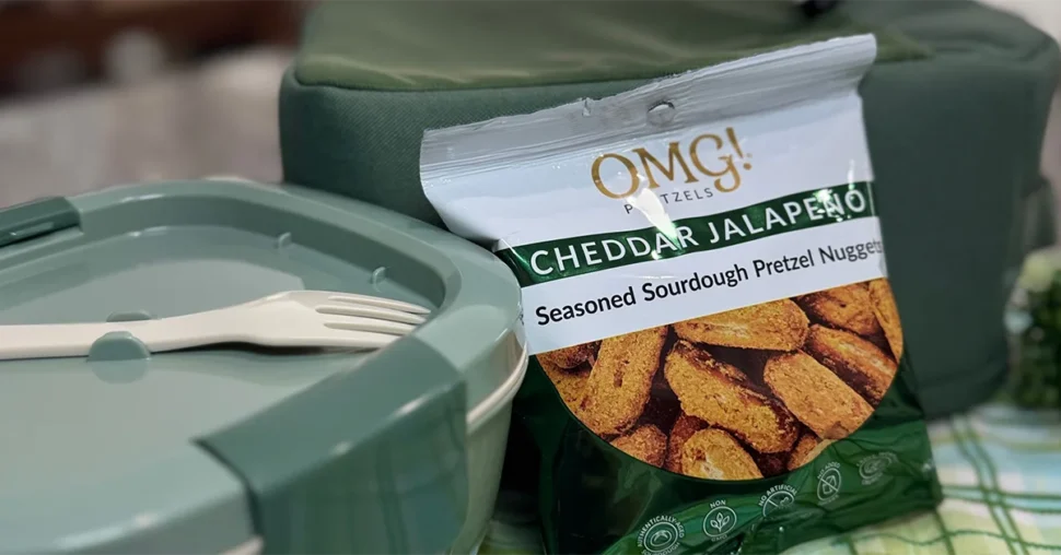 A close-up of a green lunchbox with a white spork and a bag of cheddar jalapeño sourdough pretzels. The setting feels casual and appetizing.