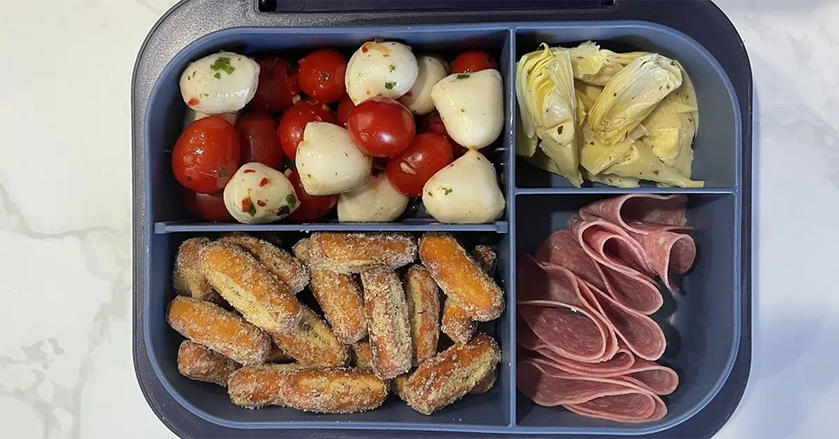 A bento box with mozzarella balls and cherry tomatoes, artichoke hearts, breaded sticks, and slices of salami. The arrangement is colorful and appetizing.