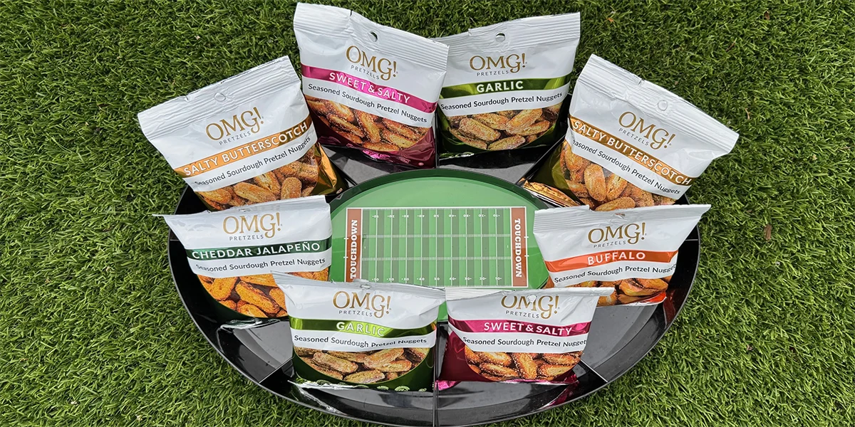 Bags of OMG! Pretzels in various flavors are arranged around a football field tray on a grass surface, creating a playful snack display.