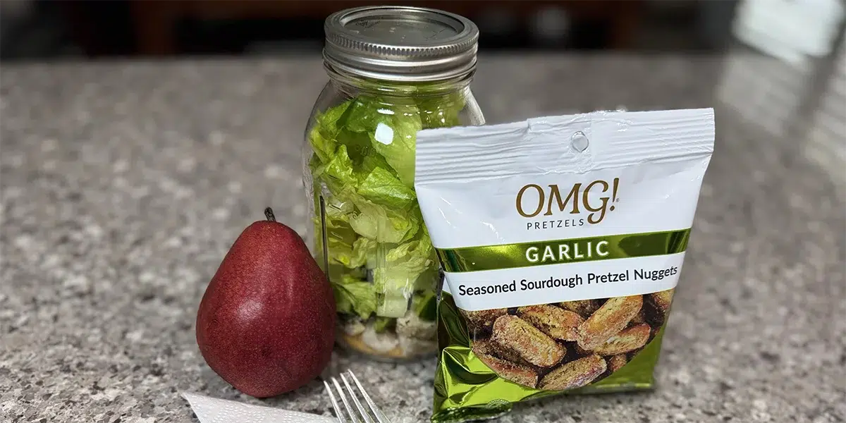 A red pear, a salad in a mason jar, and a bag of garlic pretzel nuggets are arranged on a granite countertop, evoking a fresh, crunchy snack mood.