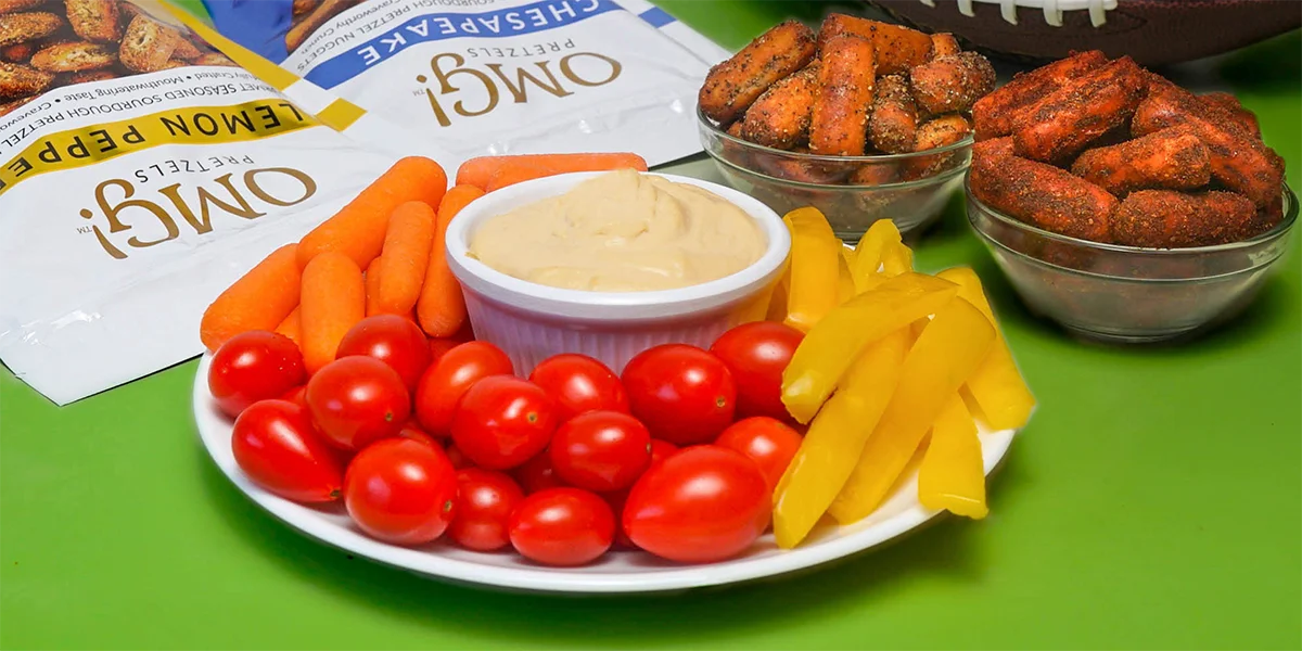 A platter with cherry tomatoes, carrot sticks, yellow pepper slices, and dip. Two bowls of seasoned pretzels and pretzel bags are visible on a green surface.