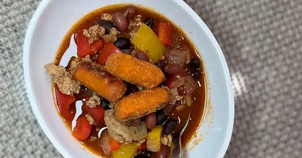 A bowl of chili with beans, ground meat, and peppers is topped with breaded fish sticks. The white bowl contrasts with a textured gray surface.