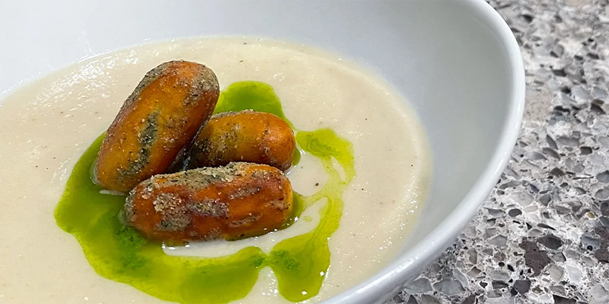 A white bowl with creamy beige soup topped with three breaded sausage pieces, drizzled with green oil. The setting includes a speckled countertop.