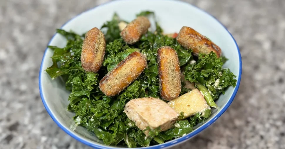 A bowl of vibrant kale salad topped with crispy potato croquettes and chunks of grilled chicken, set on a speckled gray countertop, conveying a fresh and hearty meal.
