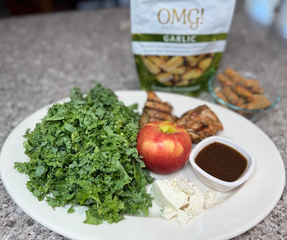 Plate with fresh kale, an apple, crumbled feta, grilled chicken, and dressing. Garlic snack bag in background suggests a healthy meal.