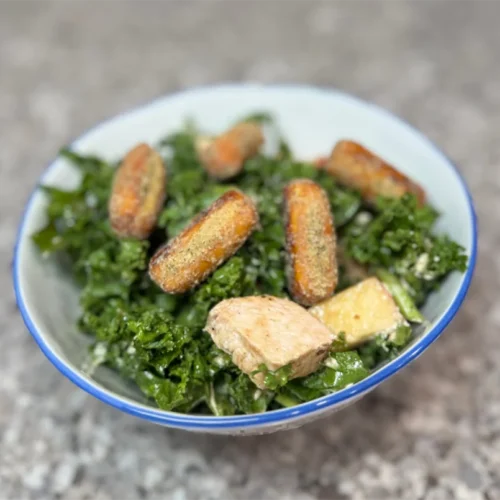 A bowl of kale salad topped with roasted potato wedges and grilled chicken on a speckled countertop. The dish appears fresh and savory.