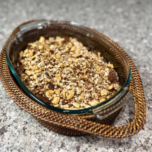 A glass baking dish filled with a crumbly oat and nut topping on a countertop. It rests in a woven rattan holder, creating a cozy, rustic feel.