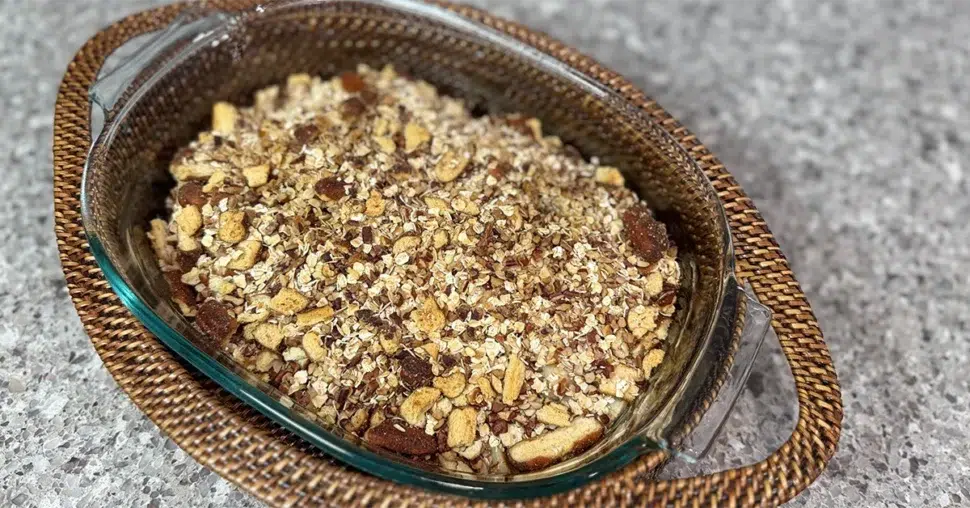 A glass bowl filled with a mixed nut and oat granola sits on a woven rattan tray atop a speckled gray countertop, conveying a rustic, homemade vibe.