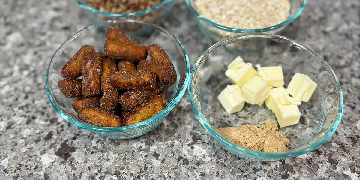 Glass bowls on a speckled countertop hold caramelized mini sausages, chopped nuts, oats, butter cubes, and brown sugar, suggesting baking ingredients.