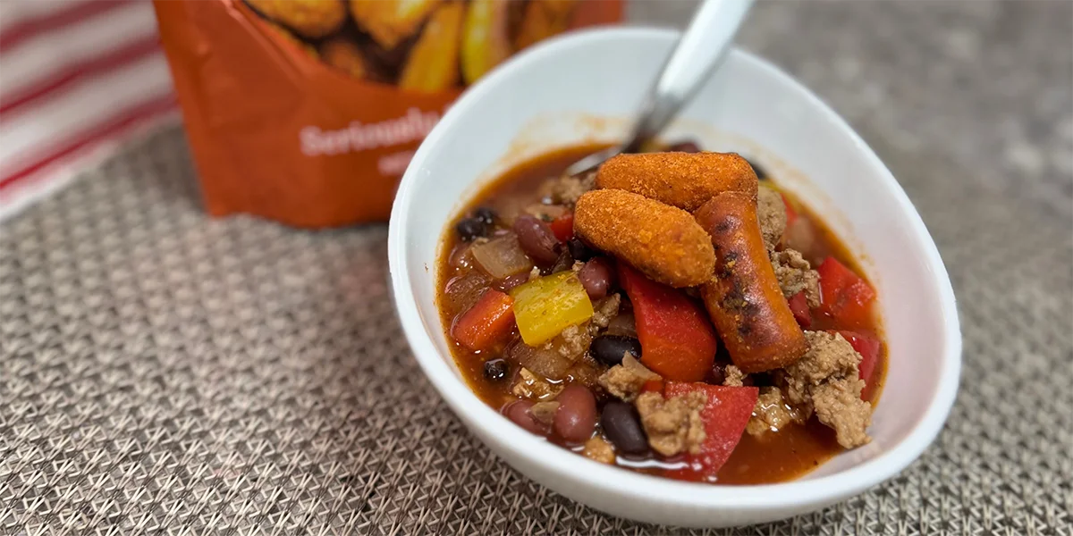 A close-up of a bowl of chili with ground meat, beans, red and yellow bell peppers, topped with crispy golden sticks. A spoon is in the bowl, and a red-striped cloth is in the background.