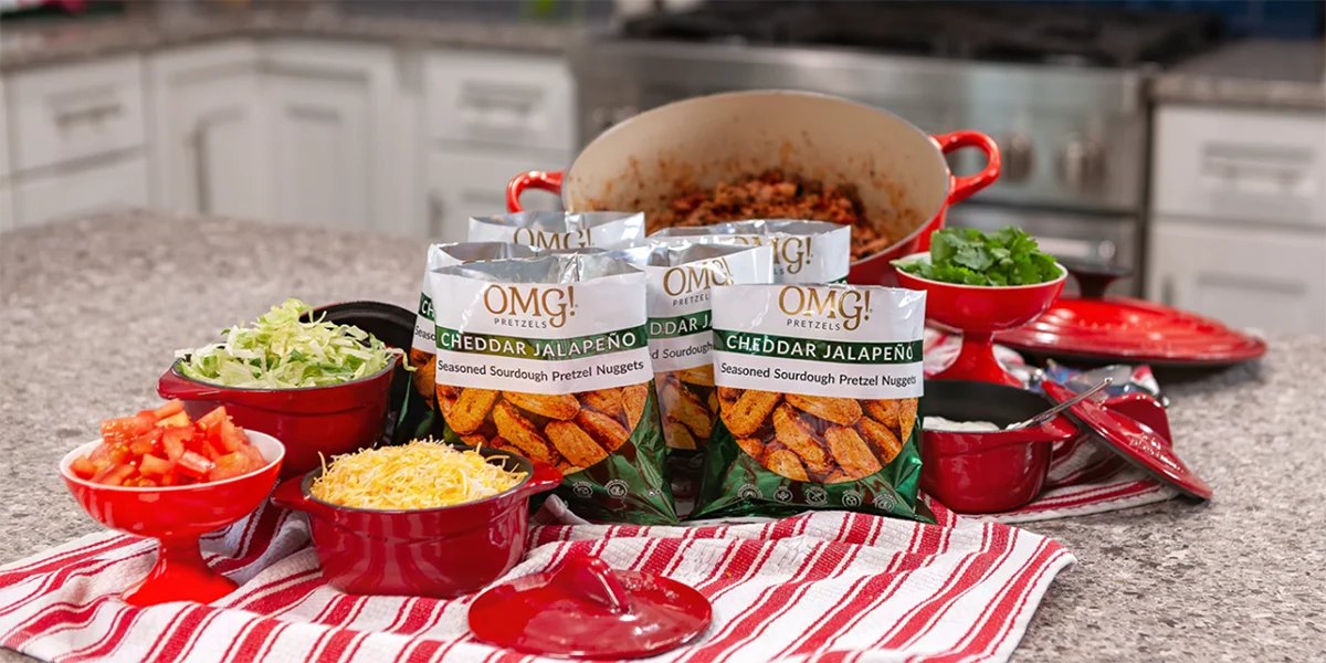 Four bags of cheddar jalapeño pretzel nuggets are on a kitchen counter surrounded by bowls of chopped lettuce, tomatoes, cheese, and cilantro on a striped cloth.